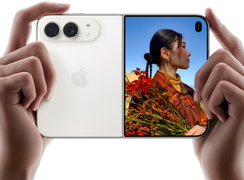 Rozevřený nebesky bílý iPhone Duo, dvě ruce ho drží na šířku, na levém panelu je vidět dvojitá 48Mpx Fusion zadní fotosoustava a vpravo se na vnějším displeji zobrazuje osoba, která se nechává fotit a díky funkci Duo Preview vidí při focení svůj obličej