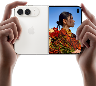 Rozevřený nebesky bílý iPhone Duo, dvě ruce ho drží na šířku, na levém panelu je vidět dvojitá 48Mpx Fusion zadní fotosoustava a vpravo se na vnějším displeji zobrazuje osoba, která se nechává fotit a díky funkci Duo Preview vidí při focení svůj obličej