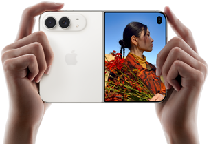 Rozevřený nebesky bílý iPhone Duo, dvě ruce ho drží na šířku, na levém panelu je vidět dvojitá 48Mpx Fusion zadní fotosoustava a vpravo se na vnějším displeji zobrazuje osoba, která se nechává fotit a díky funkci Duo Preview vidí při focení svůj obličej
